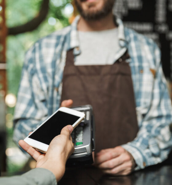 Homem barista com avental e camisa xadrez segunda máquina de cartão para cliente aproximar celular; mastercard