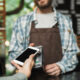 Homem barista com avental e camisa xadrez segunda máquina de cartão para cliente aproximar celular