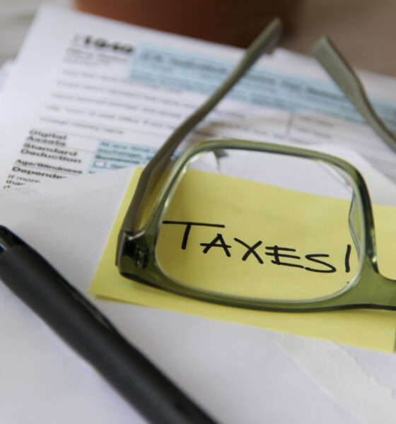 Papel com post-it amarelo escrito "Taxes" sendo ampliado por óculos e lápis perto; alíquota da CBS