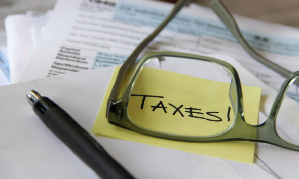 Papel com post-it amarelo escrito "Taxes" sendo ampliado por óculos e lápis perto; alíquota da CBS