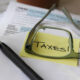 Papel com post-it amarelo escrito "Taxes" sendo ampliado por óculos e lápis perto