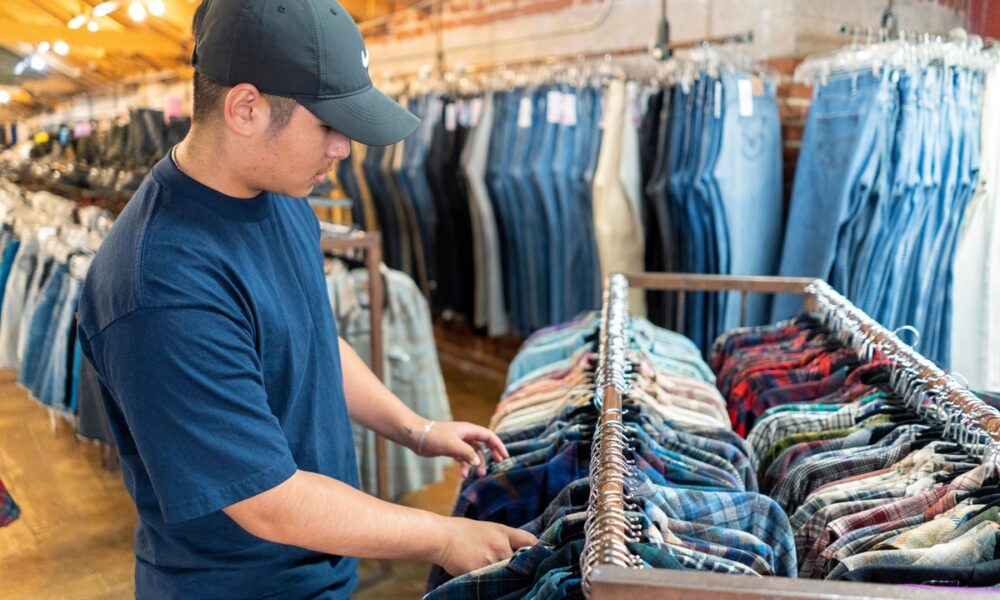 Homem de camiseta preta e boné olha camisas em arara baixa, em loja de roupas de segunda mão; lojas físicas