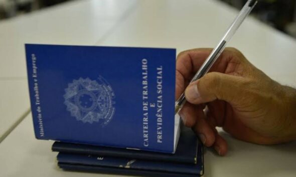Carteiras de trabalho empilhadas em uma mesa, e mão de pessoa segurando caneta assinando