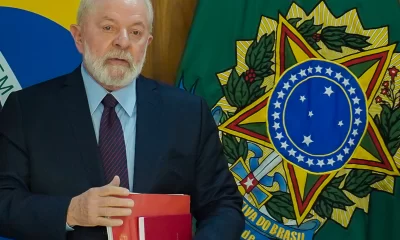 Lula com bandeiras ao fundo, de terno, segurando documentos