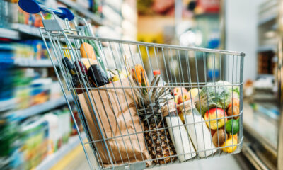 Carrinho de supermercado visto de lado, cheio de produtos como abacaxi, saco de pão, frutas. Ao fundo, prateleiras de supermercado desfocadas