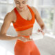 Mulher com roupa de ginástica laranja pega suplemento em pote branco; franquia de esportes