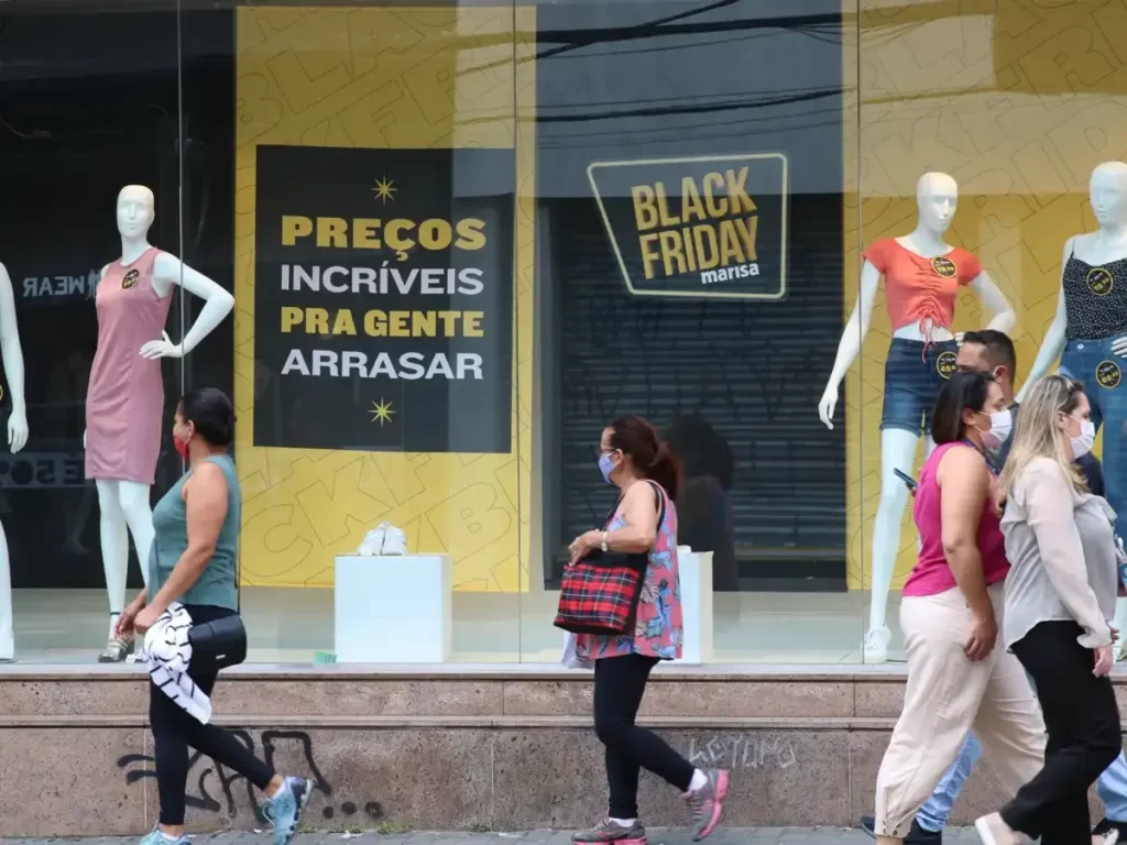 Pessoas passando em frente a loja de roupas, em promoção de Black Friday anunciada na vitrine; varejo