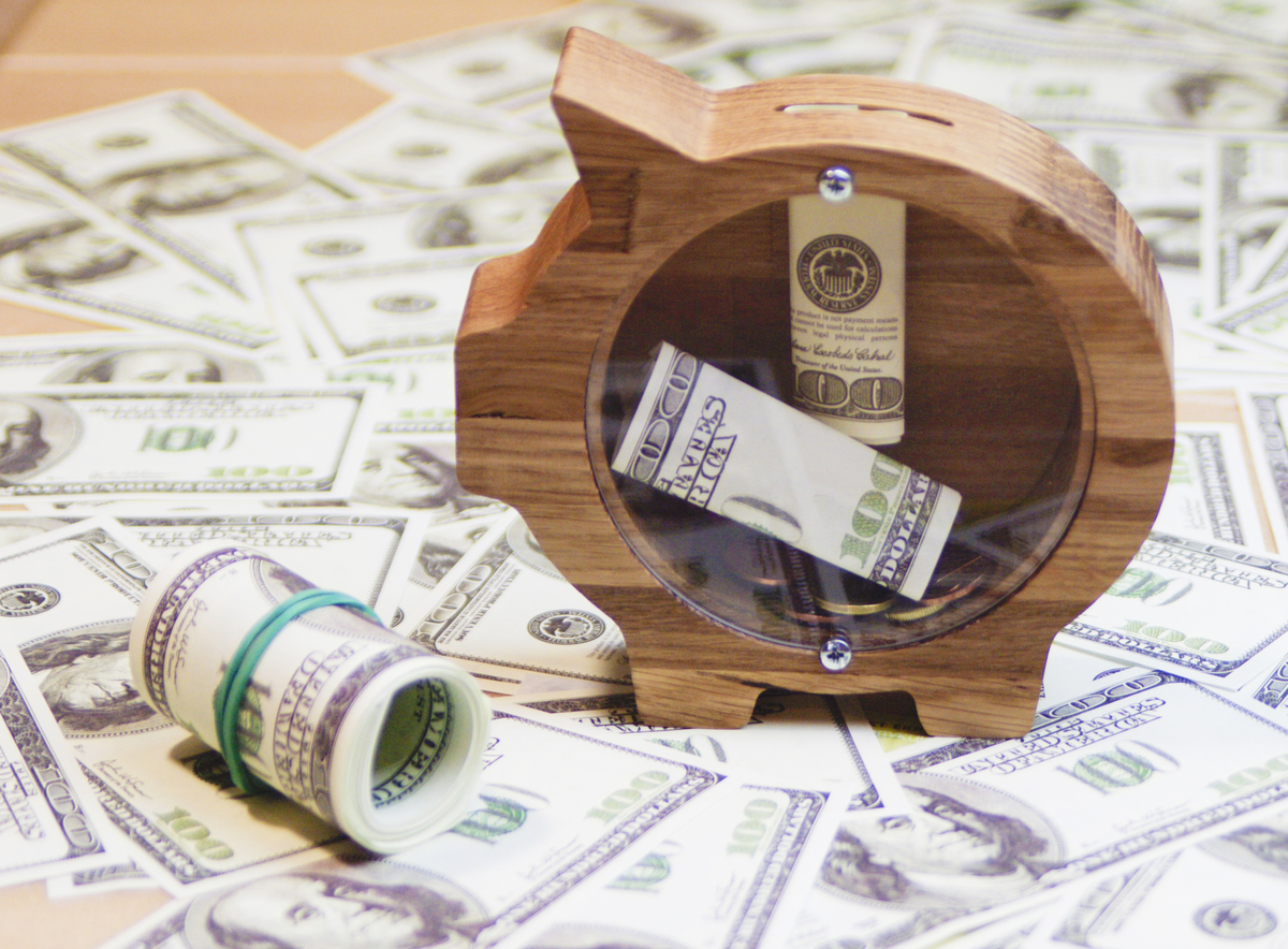 Dólares em cima de uma mesa, e um porquinho de madeira com lado de vidro e alguns dólares dentro