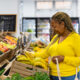 Mulher negra de blusa amarela pega bananas no supermercado, sorrindo, com sacola na mão; confiança do consumidor; deflação