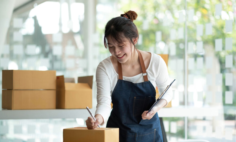 Mulher asiática organizando caixas em seu negócio D2C, usando avental azul; empresário do comércio