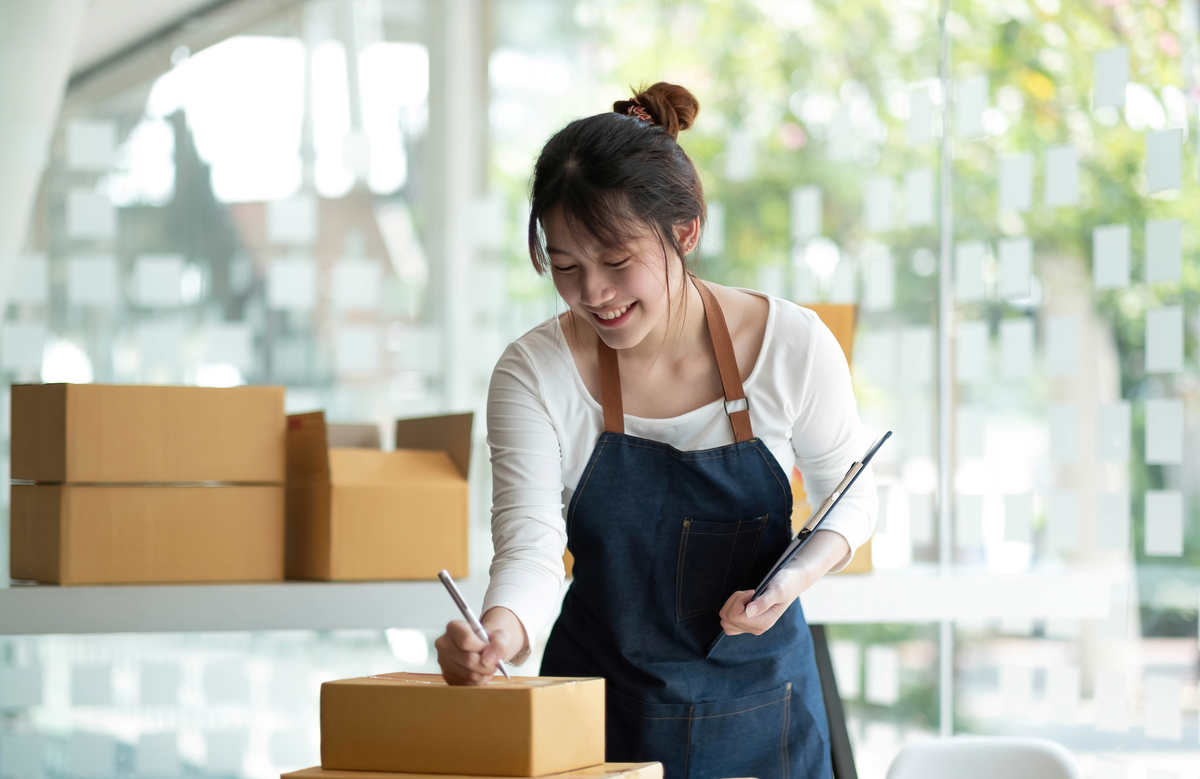 Mulher asiática organizando caixas em seu negócio D2C, usando avental azul; empresário do comércio