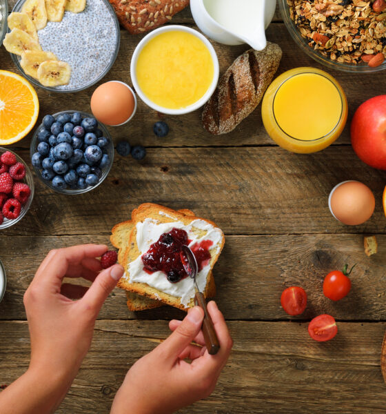 Mesa cheia de ingredientes de café da manhã, com pessoa passando geleia em torrada