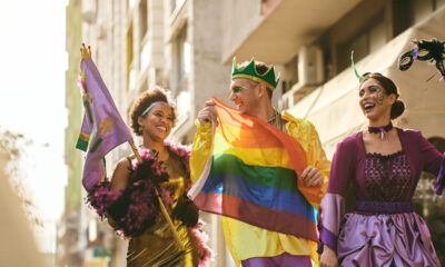 Grupo de três jovens fantasiados para o carnaval. Da esquerda para a direita são uma mulher negra com vestido dourado e segurando uma bandeira roxa; um homem com coroa verde segurando uma bandeira lgbt na frente do corpo e uma mulher com roupa roxa de princesa.