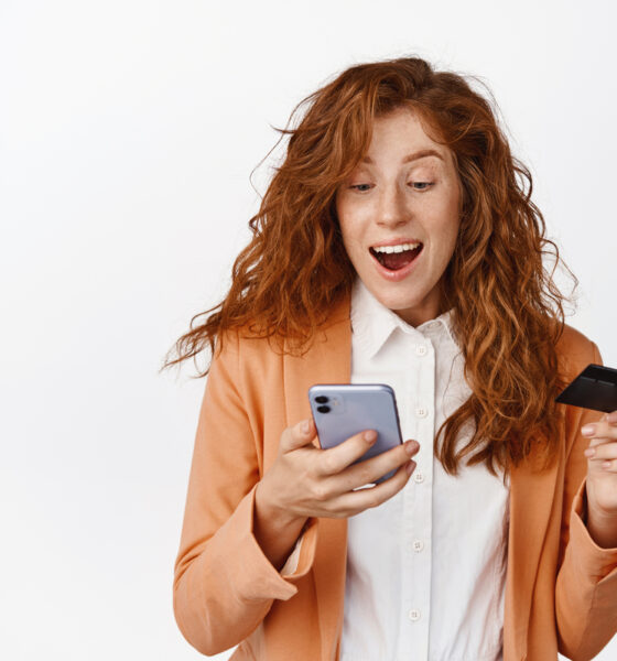 Mulher ruiva com cartão de crédito na mão olhando animada para celular; plataformas de e-commerce; varejo mobile