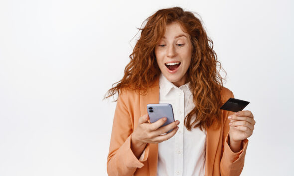 Mulher ruiva com cartão de crédito na mão olhando animada para celular; plataformas de e-commerce; varejo mobile