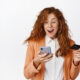 Mulher ruiva com cartão de crédito na mão olhando animada para celular; plataformas de e-commerce