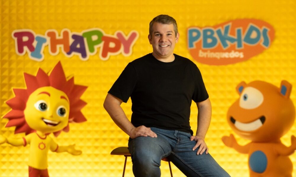 CEO da Ri Happy sentado em banquinho à frente de fundo com logos da marca e parede amarela