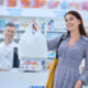Mulher sorrindo para camera com sacola de compras na farmácia. Atrás, vendedor da farmácia sorrindo.