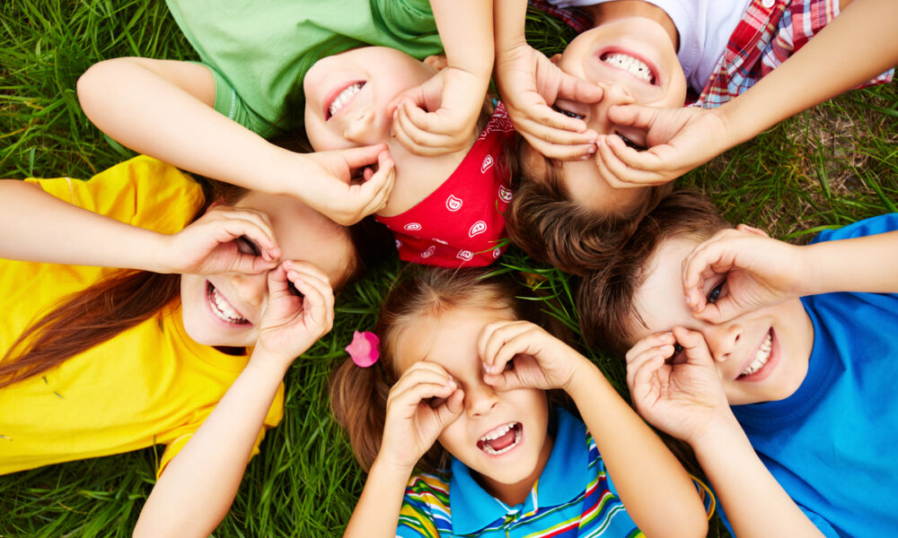 Cinco crianças deitadas na grama, sorrindo e fazendo óculos com as mãos nos olhos.